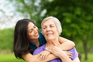 Young caregiver hugging a senior woman outdoors in a green park, both smiling.