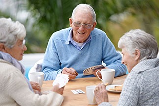 Three seniors enjoying coffee and playing cards outdoors at a senior living community.