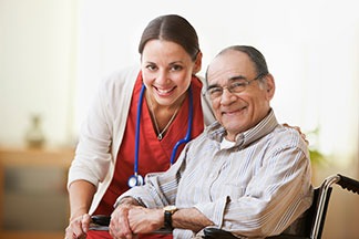 Smiling caregiver in red uniform with hand on shoulder of a senior man sitting in a wheelchair.