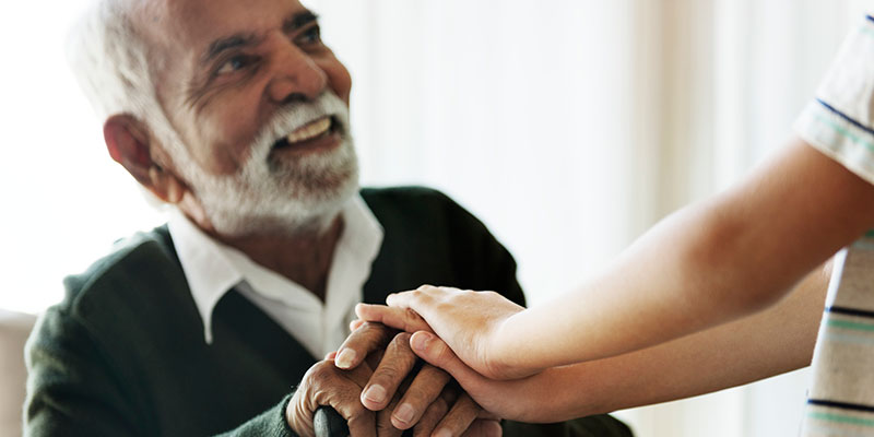 Elderly man smiling and holding hands with a younger person in a comforting gesture.