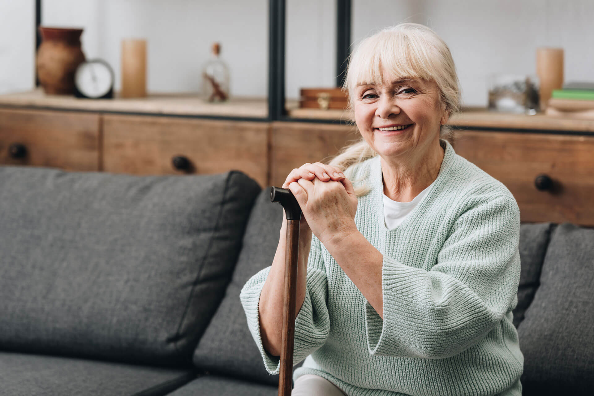 Smiling elderly woman sitting on a couch in a comfortable living space, holding a walking cane.