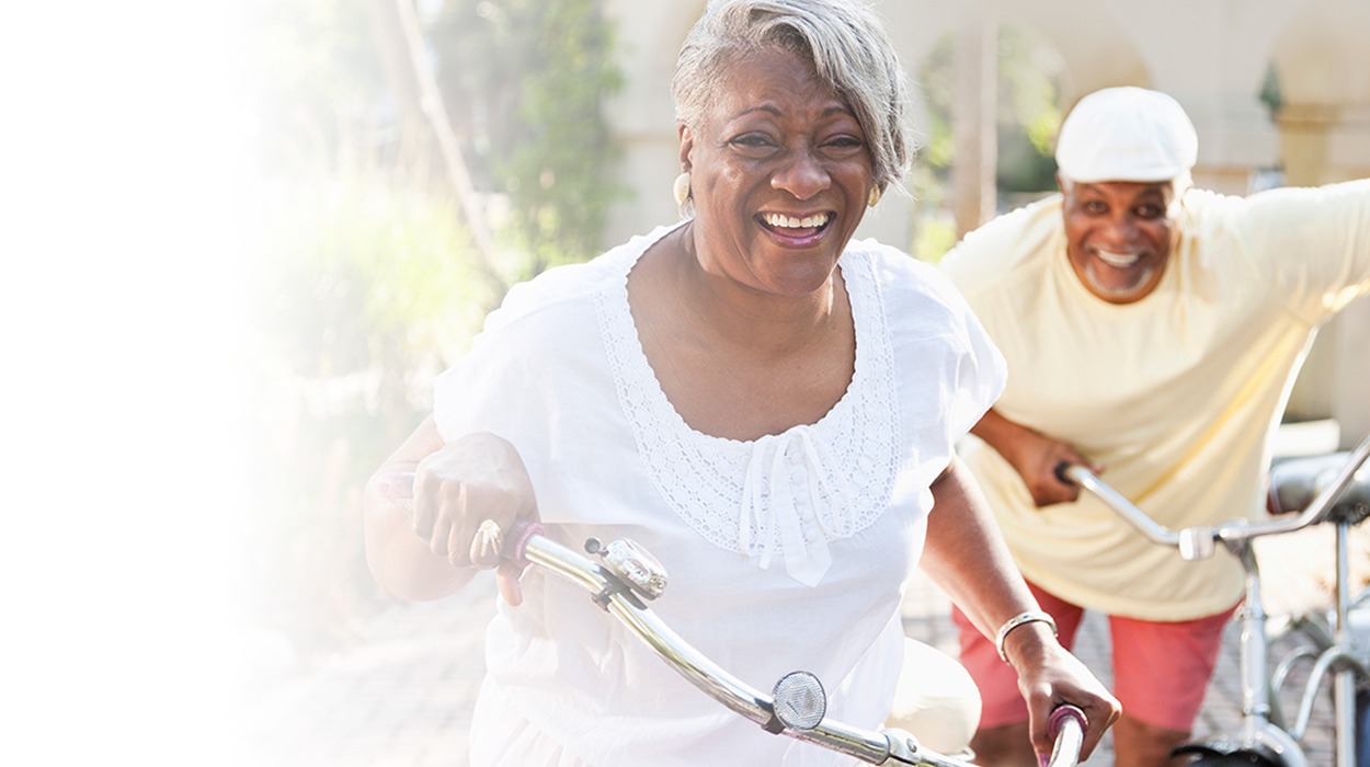 Two seniors riding bicycles and smiling outdoors at an independent living community.