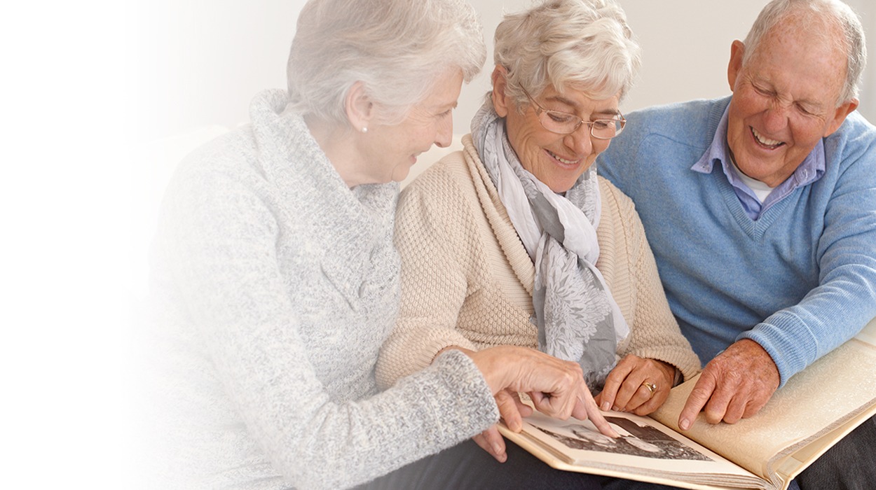Three elderly individuals sharing memories while looking at a photo album in a bright living room.