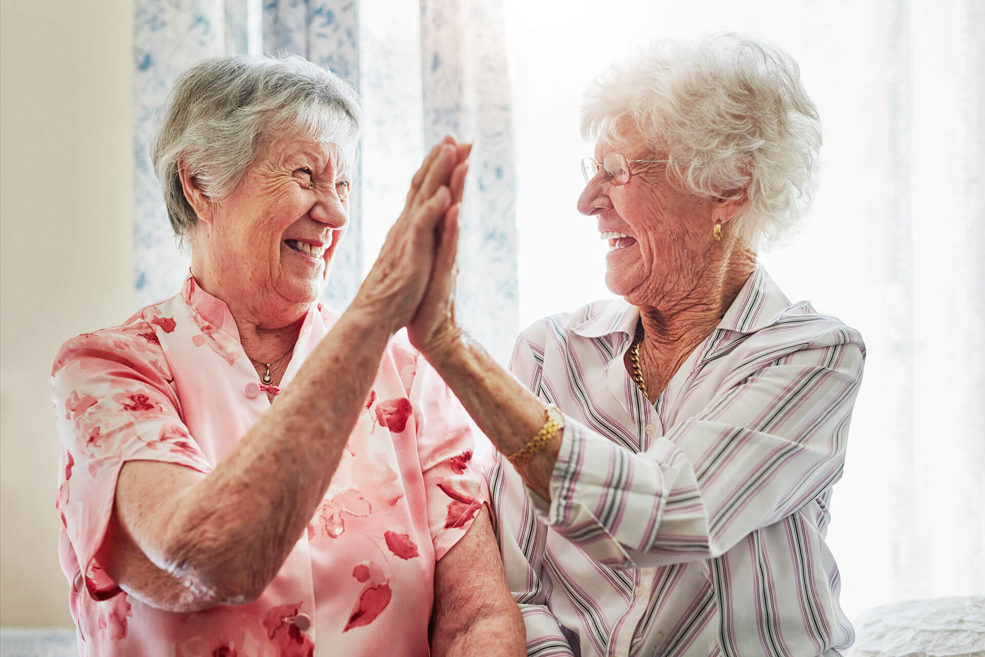 Two elderly women smiling and high-fiving in a bright room with floral curtains.