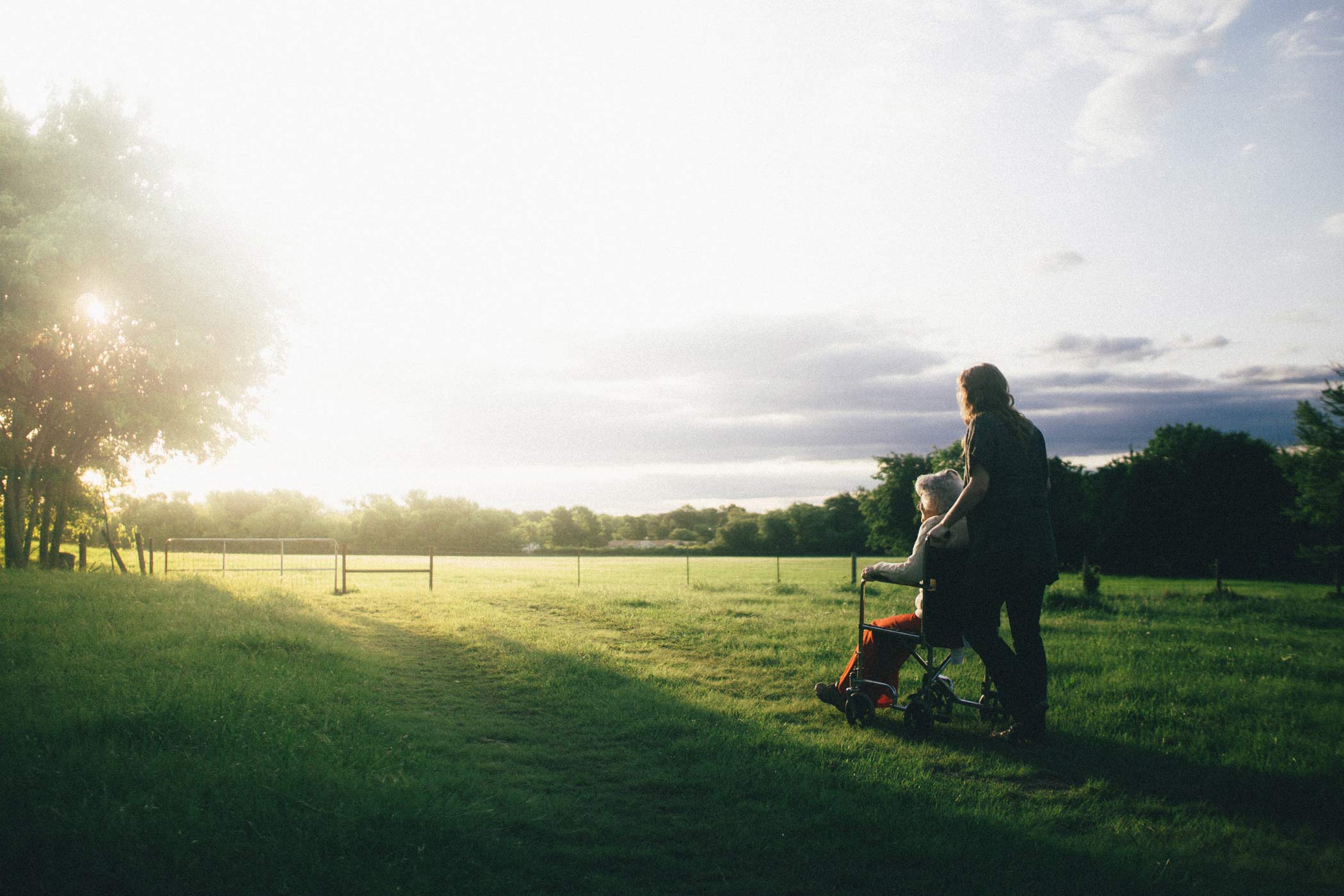 Caregiver pushing a senior in a wheelchair through a serene field at sunset.