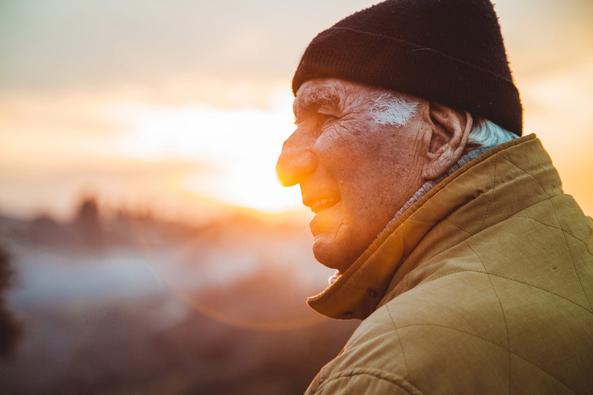 Elderly man in a beanie and coat smiling while watching the sunset outdoors.