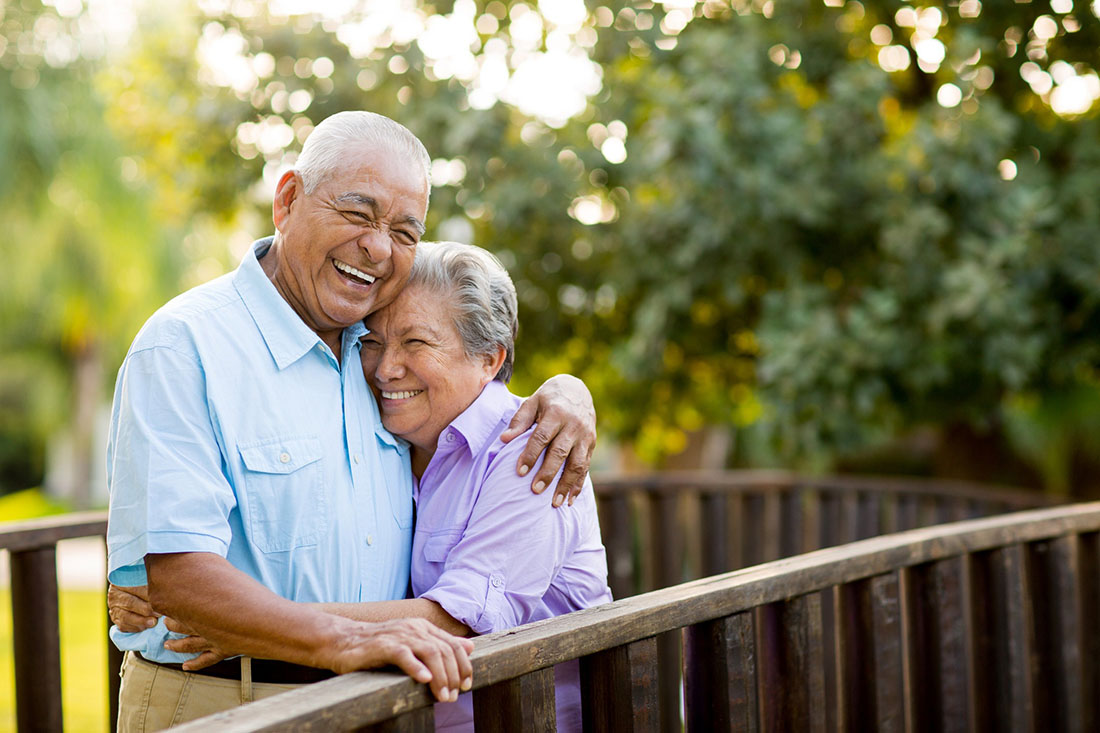 Happy senior couple embracing on a wooden bridge in a lush, green outdoor area.