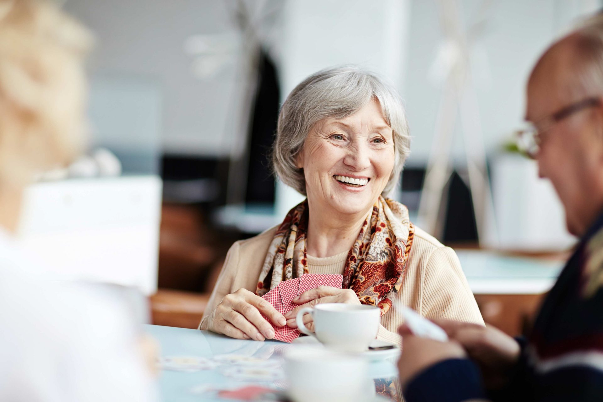 Elderly woman smiling and playing cards with friends, while enjoying a cup of coffee.