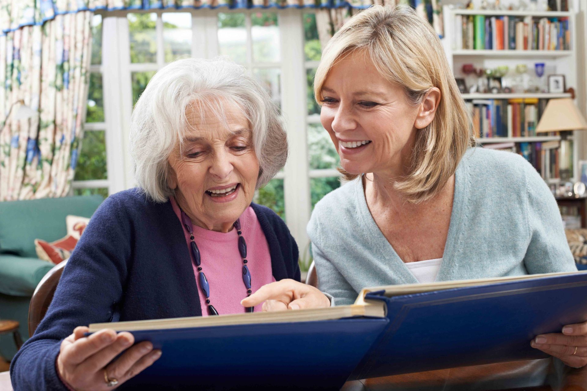 Two women smiling and looking at a photo album in a cozy living room with bookshelves.