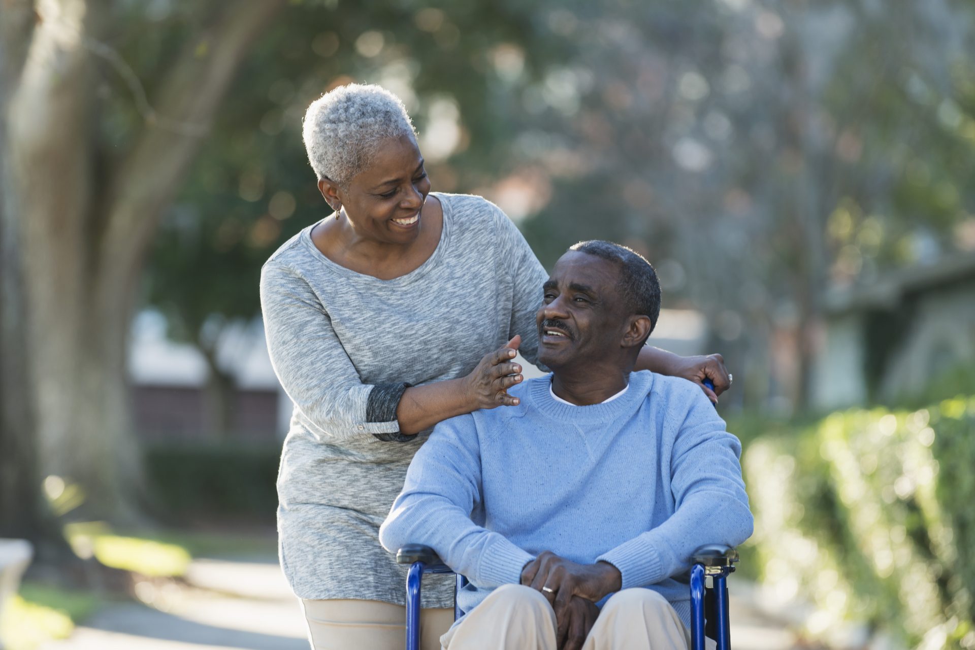 Smiling woman standing behind a man in a wheelchair outdoors at a senior living community.