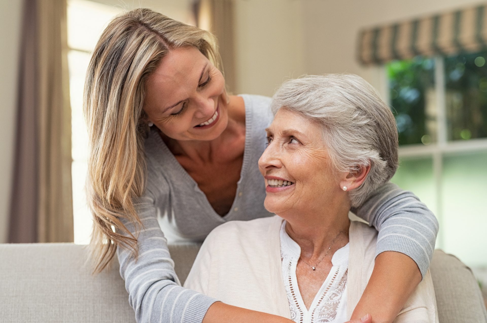 Younger woman embracing an older woman, both smiling, inside a living space.