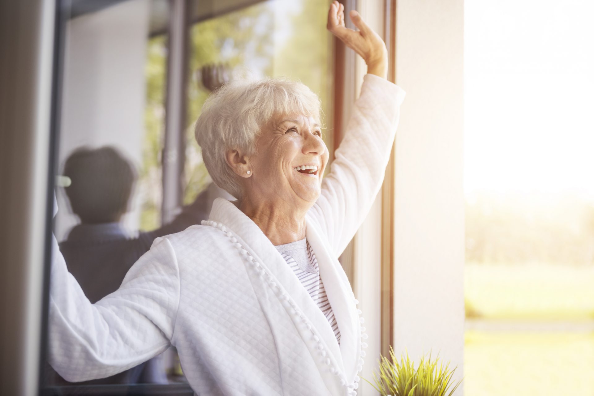 Elderly woman smiling and raising her arm while looking out of a sunny window in a unit.
