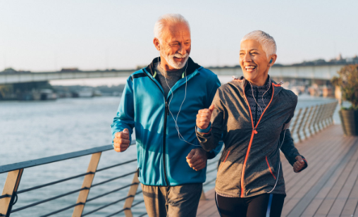 Two seniors jogging on a riverside boardwalk, listening to music on a sunny day.