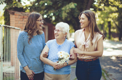 Elderly woman with flowers walking outdoors with two younger women, all are smiling.
