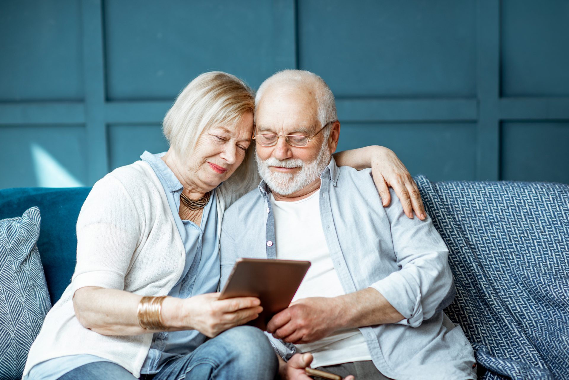 Senior couple sitting close on a couch, looking at a tablet screen with gentle smiles.