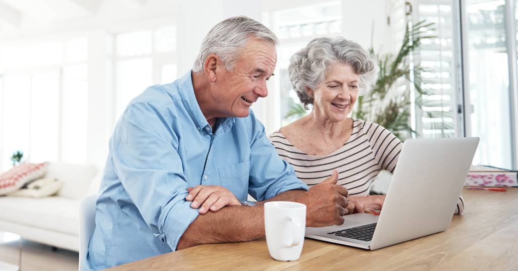 Elderly couple sitting at a table, smiling and using a laptop in their bright living space.