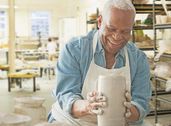 Senior man happily shaping clay pottery in a well-lit communal art room with shelves and tables.