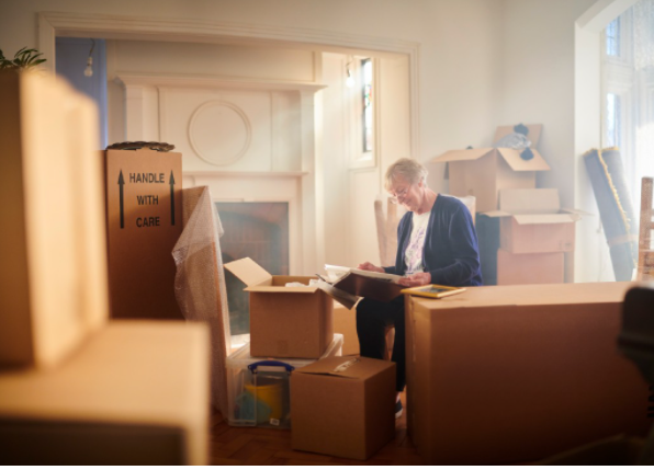 Elderly woman sitting amid moving boxes in a bright room, sorting through a large folder.