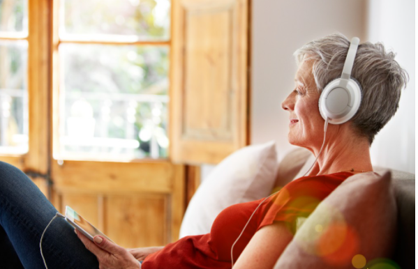 Elderly woman with headphones enjoys music in bright, cozy living room with wooden windows.