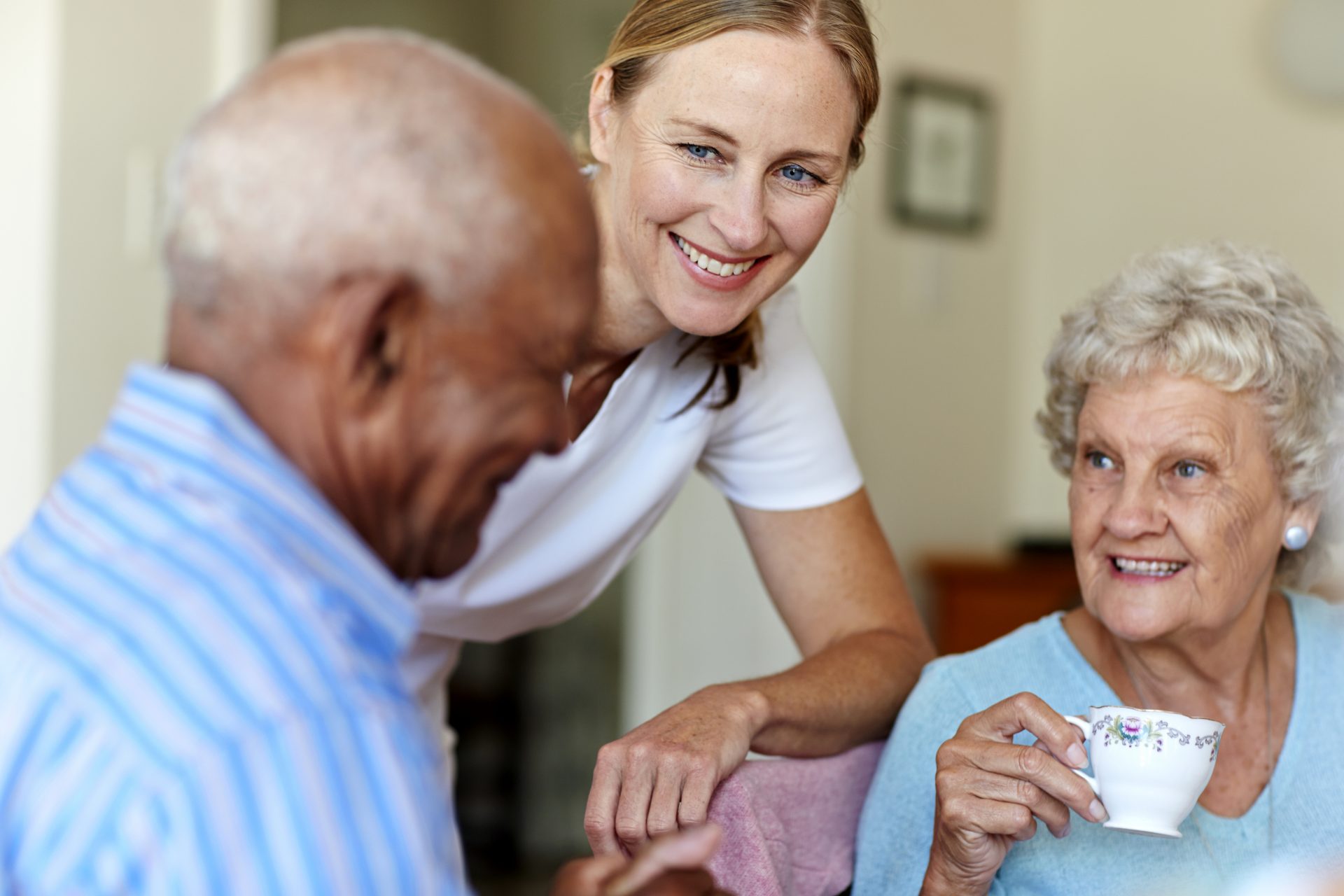 Caregiver laughing with elderly residents; one holding a teacup, the other engaged in conversation.