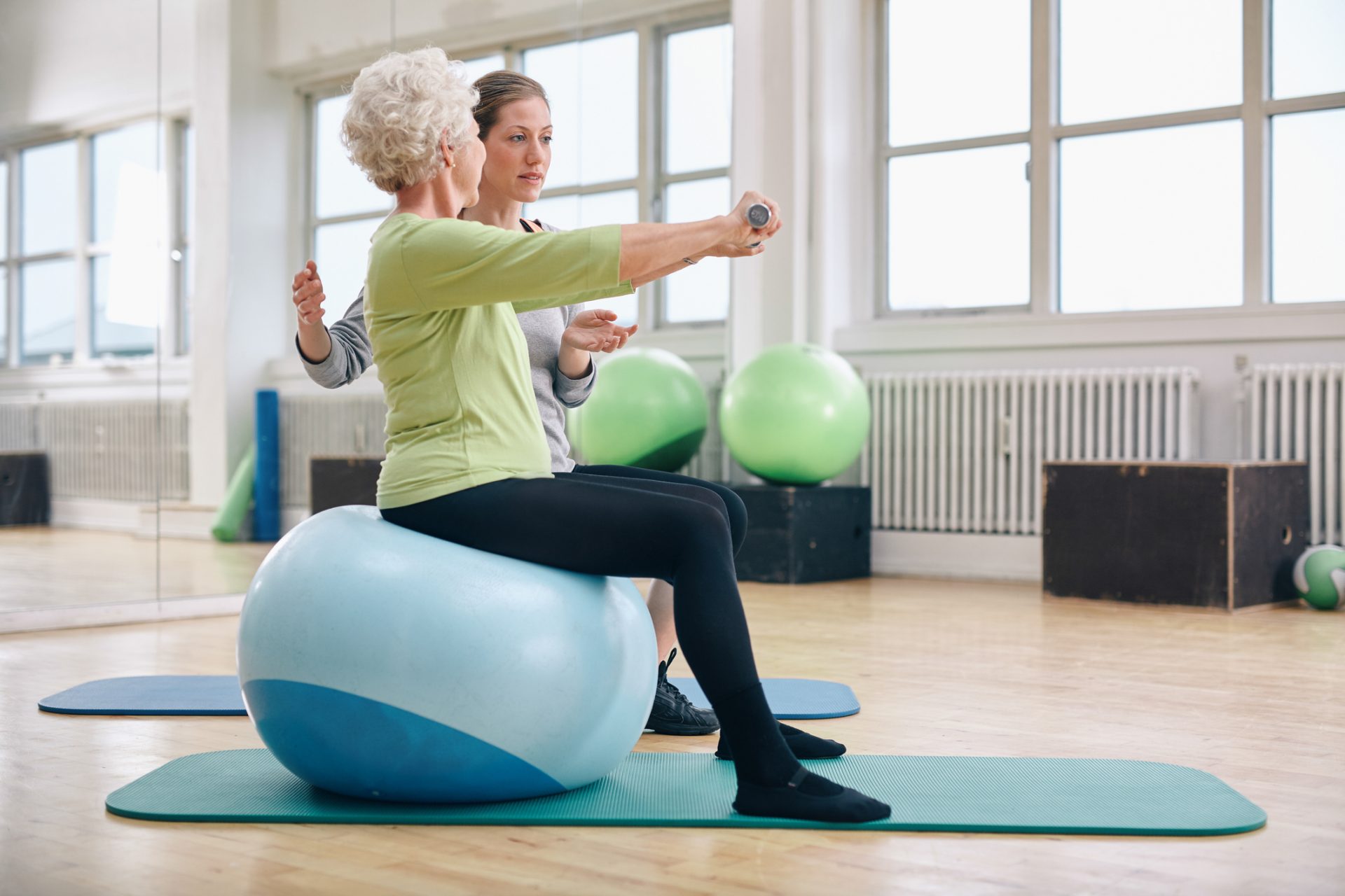 Senior woman exercising with a therapist on a balance ball in a gym with large windows.