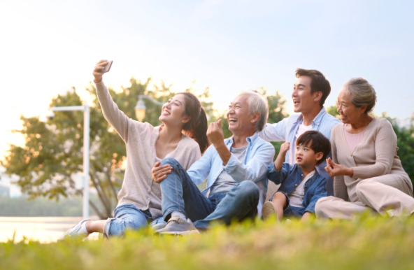 Family of five sitting on grass, smiling, and taking a selfie outdoors.