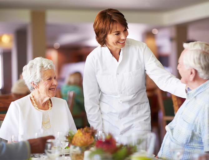 Staff member engaged with smiling elderly residents in a dining area of a senior living unit.