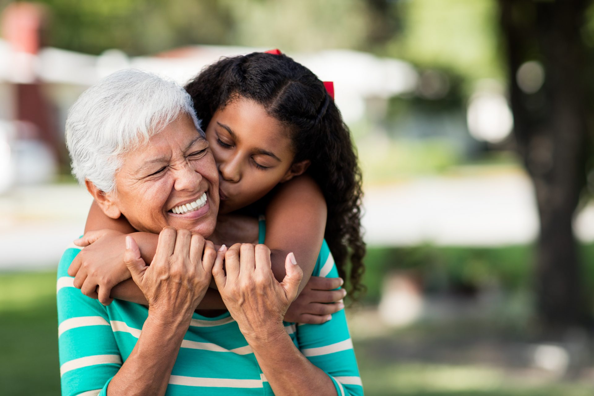 Smiling senior woman embraced from behind by young girl outdoors in a sunny community area.