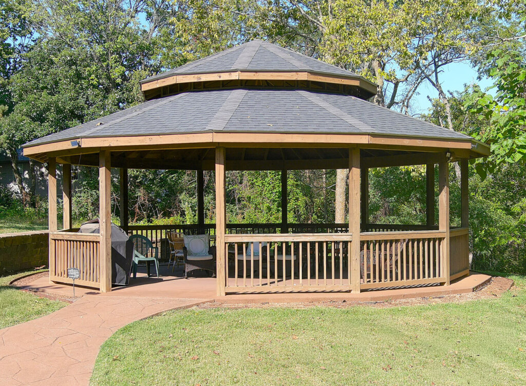 Wooden gazebo with seating area in a garden setting, surrounded by trees.